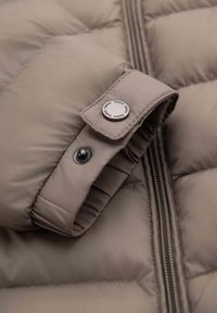 Pufferjack in lichtbeige, met een ritssluiting, een geborduurde structuur en een drukknoopaccent op de manchet voor extra detail.