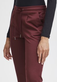 Burgundy sweatpants with a drawstring waist, side pockets, and a smooth texture; fitted design around the waist with gathered detail.