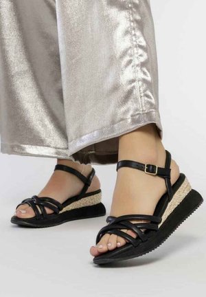 Sandalia negra con tiras entrelazadas, cuña estilo espadrille con acento de yute trenzado, suela de goma y correa ajustable en el tobillo.