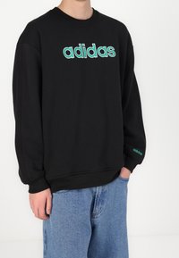 Person som bär en svart Adidas sweatshirt med turkos logotyp på bröstet och ärmarna, tillsammans med blå jeans, stående mot en enfärgad vit vägg.