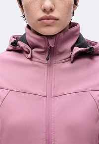 Gros plan d'une personne portant une veste rose zippée avec une doublure intérieure noire et des cordons réglables à la capuche.