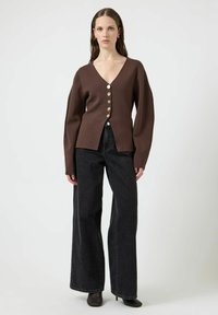 Brązowy kardigan z długim rękawem i dekoltem w serek, z złotymi guzikami z przodu, zestawiony z czarnymi spodniami dżinsowymi o wysokim stanie i szerokich nogawkach. Teksturowany materiał.