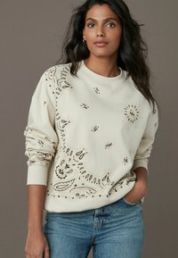 Cream sweatshirt with decorative brown paisley patterns. Features long sleeves and a crew neckline. Paired with blue jeans.