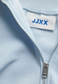 Svetlo modra jakna na zadrgo iz mehkega blaga, z originalnim srebrnim zadrgo in oznako z "JJXX" v modrem tisku.