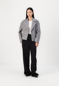 Jachetă gri scurtă, cu două rânduri de nasturi aurii, purtată peste un top alb din tricot cu textură ribbed și pantaloni negri largi, asortată cu pantofi negri.