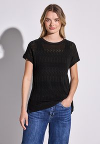 Svart stickad t-shirt med korta ärmar, rund halsringning och ett öppet vävmönster med zigzag och randiga detaljer. Kombinerad med blå jeans.