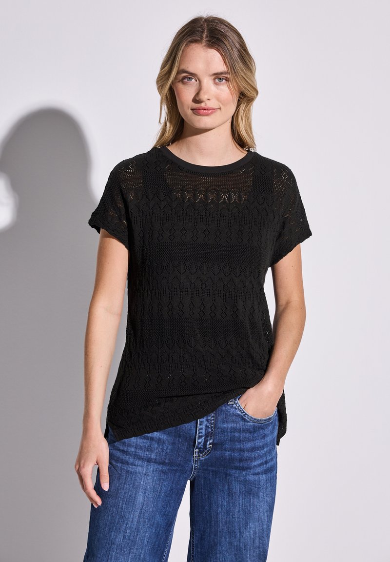 Svart stickad t-shirt med korta ärmar, rund halsringning och ett öppet vävmönster med zigzag och randiga detaljer. Kombinerad med blå jeans.