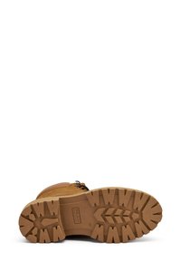 Bota de cuero marrón con suela de goma texturizada, que presenta un patrón de tacos para tracción y ojales de metal para los cordones. Logo estampado en la suela.