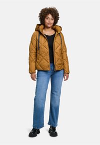 Brun foret quilted puffer-jakke med hætte, med snore, sort top, blå lige jeans og sorte platform-sko.