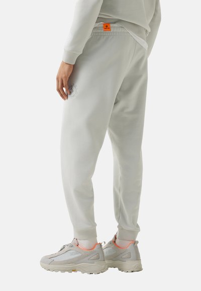 Persoon met lichtgrijze joggingbroek met geribbelde manchetten en witte sneakers met oranje accenten, staand tegen een effen witte achtergrond.
