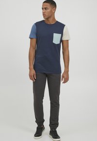 Navy blue t-shirt with a light blue pocket and multi-colored sleeves. Paired with dark gray tapered pants and black sneakers.