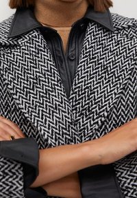 Manteau pied-de-poule noir et blanc avec un large col, superposé à une chemise noire boutonnée. Le tissu semble texturé, avec des accents en cuir lisse.