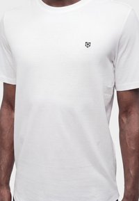 Άνδρας που φοράει λευκό απλό κοντομάνικο T-shirt με μικρό μαύρο γεωμετρικό λογότυπο στο αριστερό μέρος του στήθους, εμφανιζόμενος από τους ώμους έως τους γοφούς.