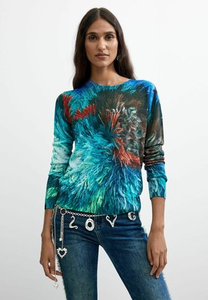 Vrouw met lang donker haar draagt een kleurrijke blauw, groen en rood patroon lange mouwen top en blauwe spijkerbroek met een zilveren bedelriem waarop "LOVE" staat.