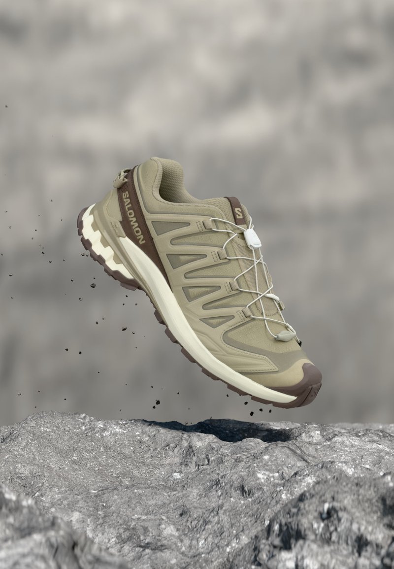 Μπεζ και καφέ παπούτσι τρεξίματος Salomon για μονοπάτια, κρεμασμένο πάνω από βραχώδες έδαφος με μικρά κομμάτια να πέφτουν γύρω του.