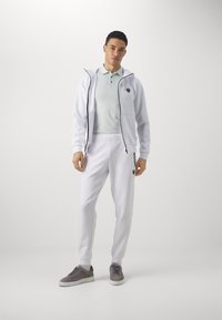 White zip-up hoodie with black accents, paired with white joggers and grey sneakers. Smooth fabric, fitted design, and logo detail on chest.