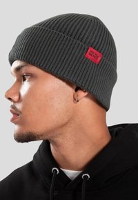 Mladý muž v černé mikině s kapucí, nosící tmavě šedou pletenou čepici s červeným nápisem "WRONG FRIENDS", čelící vlevo na šedém pozadí.