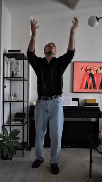 Homem em camisa preta e jeans a lançar uma almofada cinza numa sala de estar moderna com prateleira preta, piano, planta e obras de arte emolduradas.