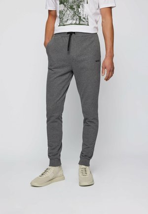 Mężczyzna w szarych dresowych spodniach z czarnym sznurkiem, białych trampkach i białej koszulce z nadrukiem, stojący na jednolitym jasnym tle.