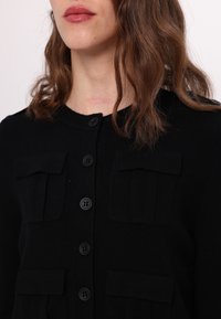 Femme aux cheveux bruns mi-longs portant un cardigan noir boutonné avec quatre poches à rabat, lèvres légèrement brillantes.