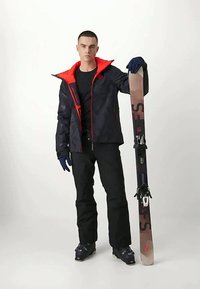 Veste de ski noire avec des accents rouges, pantalon noir, gants et chaussures de ski. Tenant un ski avec une base dégradée et des fixations.