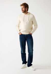 Beež kapuutsiga dressipluus reljeefse logoga ja paeladega, koos tumesiniste teksade ja valgete tennistega. Modell seisab neutraalsel taustal.