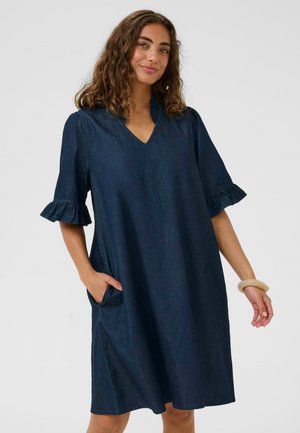 Nő térdig érő sötétkék farmerruhában, V-nyakkivágással, fodros könyökhosszú ujjakkal és oldalzsebekkel, zsebébe tett kézzel mosolyog.