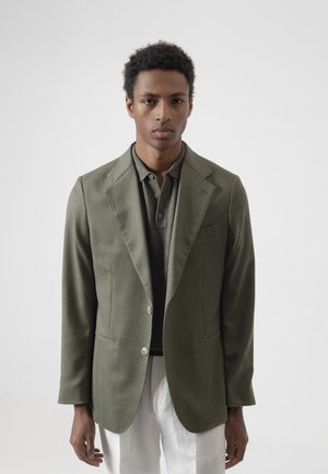 Joven con cabello corto y rizado que lleva una chaqueta verde oliva sobre una camiseta tipo polo oscura y pantalones blancos, de pie frente a un fondo claro y liso.