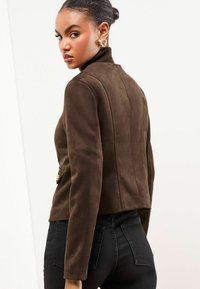 Veste en suede marron avec un col haut, une coupe ajustée et des coutures verticales, ornée d'un bouton décoratif en métal doré à la taille.