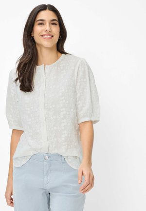 Frau mit langen dunklen Haaren lächelt, trägt eine weiße, kurzärmelige Bluse mit Knöpfen und floralem Muster sowie hellblaue Hosen vor einem weißen Hintergrund.