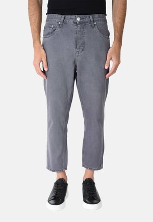 Uomo che indossa jeans grigi corti e sneaker nere con suola bianca che sta in piedi davanti a uno sfondo semplice.