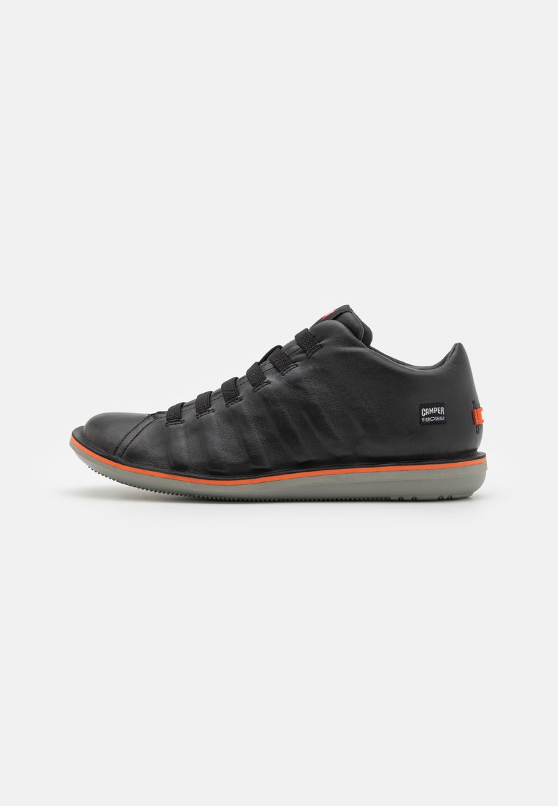 Black leather shoe with textured side panel, elastic laces, orange accent on sole, and a logo on the heel. Slim, rounded silhouette.