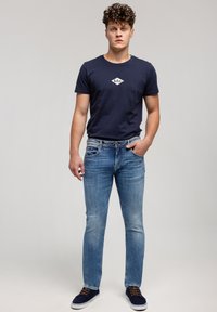 Młody mężczyzna z kręconymi włosami, ubrany w granatową koszulkę z logo, jasnoniebieskie dżinsy i granatowe trampki, stoi z jedną ręką w kieszeni.