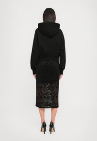 Kvinne står med ryggen til, iført en svart kort hoodie, svart blomsterlaced midi-skjørt og svarte høyhælte sko.