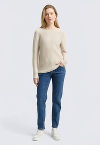 Beżowy długi sweter z długim rękawem, okrągły dekolt, luźny krój. W zestawie z niebieskimi jeansami o prostych nogawkach i białymi trampkami. Prosty i elegancki design.