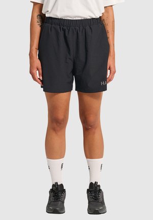Schwarze Sportshorts mit einem elastischen Bund, mit einem Logodetail am unteren linken Bein. Getragen mit weißen Socken und schwarzen Schuhen.