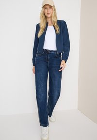 Marine bomberjack met elastische manchetten, witte t-shirt, donkerblauwe jeans met zijstrepen en witte sneakers. Lichtbeige pet maakt de look compleet.