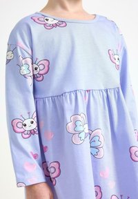 Barn klädd i en ljuslila klänning med långa ärmar, dekorerad med leende rosa och blå fjärilar samt rosa hjärtan.