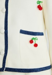 Chaqueta de textura crema con ribetes azul marino, botones cubiertos de tela, bolsillo frontal y embellishments de cereza roja con cuentas.