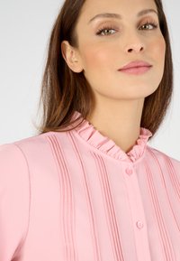 Blusa rosa con cuello con volantes, pliegues verticales y cierre de botones. La textura suave y el sutil brillo realzan su diseño.