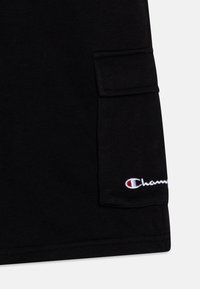 Black cotton T-shirt featuring a side pocket and embroidered "Champion" logo in white and red on the lower front. Smooth texture.
