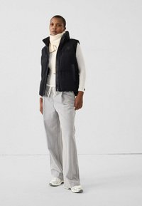 Donna che indossa un gilet imbottito nero, maglione a collo alto bianco, pantaloni grigi a gamba larga e sneakers argentate, in piedi davanti a uno sfondo bianco uniforme.