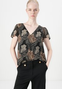 Blusa corta a maniche corte marrone e nera con scollatura a V, caratterizzata da una stampa animale dorata e motivi astratti, indossata sopra pantaloni neri con bottoni dorati.