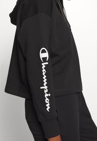 Sudadera corta negra de mezcla de algodón, con cierre frontal, puños acanalados y logo "Champion" en blanco en la manga izquierda.