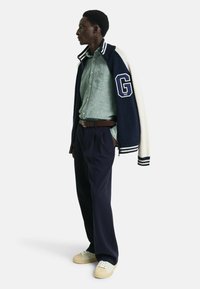 Marineblauwe en crèmekleurige varsityjack met een pluizige textuur en gestreepte manchetten, gedragen over een lichtgroen overhemd met kraag, gecombineerd met marineblauwe broek en beige sneakers.