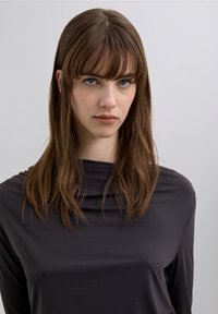 Long-sleeve top in dark gray fabric, featuring a draped neckline and smooth texture. Model has long, straight hair with bangs.