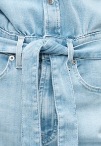 Φωτεινό γαλάζιο τζιν παντελόνι με ζώνη μπροστά σε κόμπο, ασημένιες καρφίτσες και εμφανείς ραφές γύρω από τις τσέπες και τη μέση.