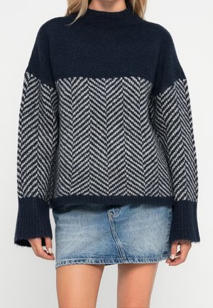Frau trägt einen marineblauen Pullover mit grauem Chevron-Muster und einem blauen Jeans-Minirock, stehend vor einem weißen Hintergrund.