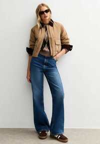 Tansfärgad bomberjacka med bruna accenter, argyletröja och blå utsvängda jeans. Bruna slip-on-skor med vita ränder, solglasögon. Neutral bakgrund.