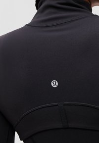 Primer plano de la parte trasera de una chaqueta deportiva negra con un pequeño logo reflectante de Lululemon centrado debajo del cuello.